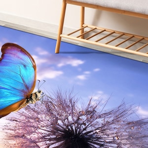 May include: A blue butterfly with brown wings lands on a purple dandelion with dew drops. The background is a blue sky with white clouds.