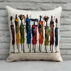 Puede incluir: Una almohada decorativa con un diseño abstracto colorido de figuras que llevan cestas en la cabeza. Las figuras están pintadas en tonos de rojo, amarillo, verde, azul y negro sobre un fondo blanco.