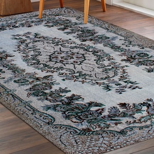 Può includere: Un tappeto bianco con un motivo floreale in tonalità di verde, marrone e beige. Il tappeto ha un medaglione centrale con un motivo ripetitivo di foglie e fiori.