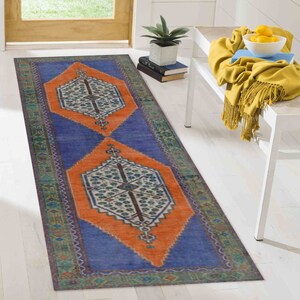 Könnte beinhalten: Ein langer, rechteckiger Teppich mit einem geometrischen Muster in Blau und Orange. Der Teppich hat einen Rand mit grünen, violetten und weißen Details. Der Teppich liegt auf einem weißen Holzboden.