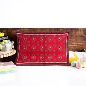Peut inclure: Un coussin rouge avec un motif géométrique, des œufs de Pâques, une figurine de lapin et un présentoir à gâteaux. Le coussin est rectangulaire avec une bordure foncée. Les œufs de Pâques sont de couleurs pastel.