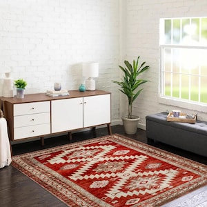 Op de afbeelding: Een rood en crèmekleurig vloerkleed met een geometrisch patroon. Het vloerkleed heeft een verweerde uitstraling en ligt op een houten vloer in een kamer met witte bakstenen muren.