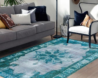 Könnte beinhalten: Ein türkisfarbener und weißer Vintage-Teppich mit einem verblassten Blumenmuster. Der Teppich liegt auf einem Holzboden in einem Wohnzimmer mit einem grauen Sofa und einem weißen Sessel.