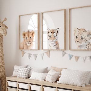 May include: Three framed watercolor paintings of baby animals. The paintings feature a lion, a tiger, and a leopard. The paintings are hung on a white wall with a wooden frame.