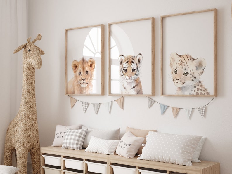 May include: Three framed watercolor paintings of baby animals. The paintings feature a lion, a tiger, and a leopard. The paintings are hung on a white wall with a wooden frame.