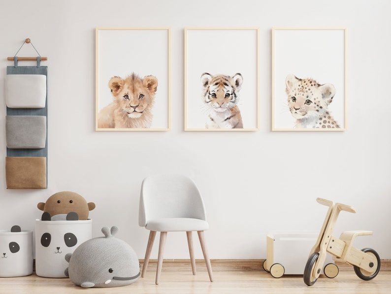 May include: Three framed watercolor paintings of baby animals. The paintings are of a lion, a tiger, and a leopard. The paintings are hung on a white wall in a nursery.