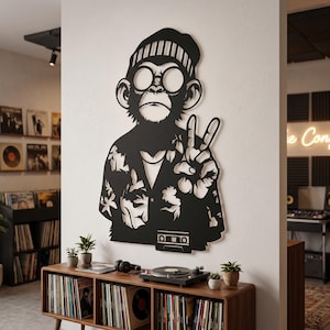 Puede incluir: Una pieza de arte mural de metal negro que representa a un mono estilizado con gorro y gafas redondas, vestido con una camisa estampada y haciendo el signo de la paz. El diseño incluye un gráfico de un casete en la parte inferior.