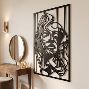 Op de afbeelding: Een rechthoekig zwart metalen wandkunstwerk met een gestileerde, doorlopende lijntekening van een menselijk gezicht met vloeiend haar, omsloten door een verticaal frame. Het ontwerp gebruikt abstracte, gebogen lijnen.