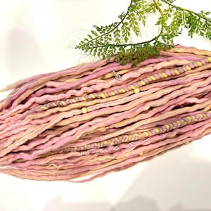 Puede incluir: Dreadlocks rosas y amarillo claro con cuentas amarillas y un anillo plateado. Las rastas están dispuestas en un haz, con algunos mechones trenzados. Una ramita de follaje verde en el fondo.