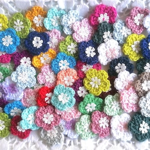 Könnte beinhalten: Eine Sammlung gehäkelter Blumenverzierungen in verschiedenen Farben, darunter Rosa, Weiß, Grün, Blau und Gelb. Die Blumen sind alle handgefertigt und haben ein zartes, filigranes Design.