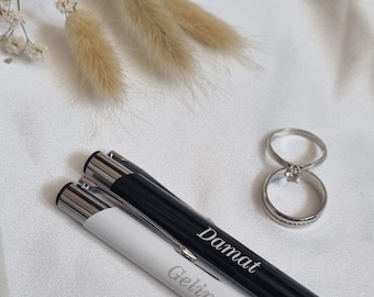 May include: Two pens with silver accents, one black and one white, with the words "Damat" and "Gelin" engraved on them. The pens are lying on a white surface with two silver rings.