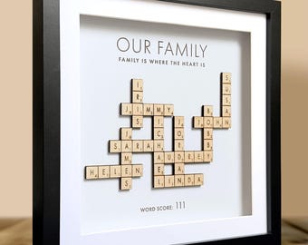 May include: A square black frame containing a white mat with the text OUR FAMILY and FAMILY IS WHERE THE HEART IS. Wooden letter tiles are arranged in a crossword pattern featuring the names Jimmy, Sarah, Helen, Audrey, Linda, John, and Susan.
