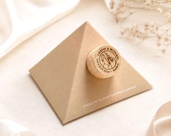 May include: A tan pyramid-shaped paper card with a circular cork topper attached to the front. The cork features a stamped illustration of two hands holding each other with the text "LOUISE & MICHAEL 24TH JULY 2025 MADRID". The card base includes the text "UNFOLD ME TO LEAVE YOUR SPECIAL MESSAGE".