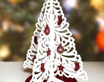 May include: A white, laser-cut Christmas tree with red glitter ornaments, including stockings, stars, and ball shapes. The tree sits on a round, red glitter base, creating a festive holiday decoration.