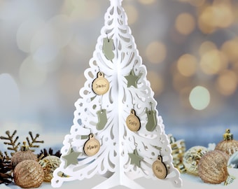 May include: White wooden Christmas tree with cutout design. The tree is decorated with wooden ornaments with names written on them, including "Jamie", "Papa", "Maria", and "Aila".