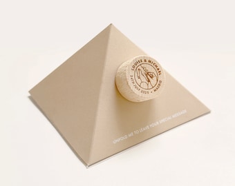 May include: A beige pyramid-shaped box with a wooden round topper. The topper has the text "LOUISE & MICHAEL 24TH JULY 2025 MADRID" engraved on it. The box has the text "UNFOLD ME TO LEAVE YOUR SPECIAL MESSAGE" printed on the bottom.