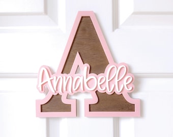 May include: A wooden letter "A" in pink with a brown center, featuring the name "Annabelle" in white script. The sign is mounted on a white door, suitable for a child's room or nursery.