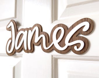 May include: A wooden name sign with the name "James" written in white cursive letters. The sign is made of dark brown wood with a natural wood grain.