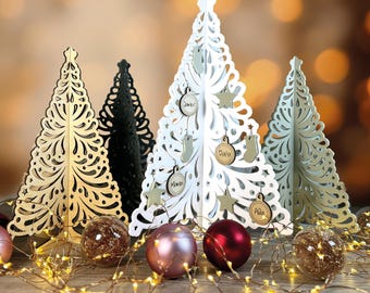 May include: Four decorative wooden Christmas trees with intricate cutouts. The trees are white, gold, black, and green. Each tree has small wooden ornaments with names written on them. The trees are surrounded by a string of fairy lights and decorative ornaments.