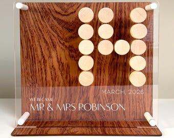 May include: A wooden wedding sign with a clear acrylic overlay. The sign features the text "WE BECAME MR & MRS ROBINSON MARCH, 2026". The wooden background has a natural wood grain pattern, and the letters are formed by small, round wooden pieces.