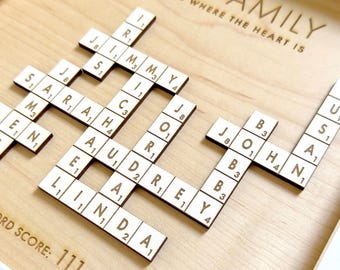 May include: A framed wooden wall art piece featuring a crossword puzzle design with light-colored wooden tiles. The text reads OUR FAMILY, FAMILY IS WHERE THE HEART IS, and WORD SCORE: 111. The tiles display names including JIMMY, SARAH, AUDREY, LINDA, JOHN, and USA.