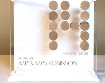 May include: A clear acrylic wedding sign with a white background. The sign features wooden circles arranged to form the letter "M". Text includes "WE BECAME", "MR & MRS ROBINSON", and "MARCH, 2026".