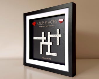 May include: A framed artwork with a black background and white matting. The text "OUR PLACES" and "OUR LOVE ADVENTURES" is displayed. White letter tiles spell out locations like New York, Athens, and Phuket. Two red heart accents are included.