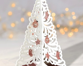May include: A white, laser-cut Christmas tree with decorative cutouts and glittery rose gold ornaments, including stars, baubles, and stockings. The tree stands on a rose gold glitter base, creating a festive holiday decoration.