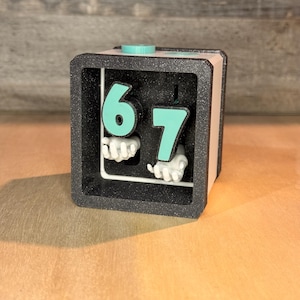 Könnte beinhalten: Ein schwarzer und beigefarbener würfelförmiger Schreibtischwecker mit den Zahlen 6 und 7 in Türkis. Die Zahlen werden von kleinen, weißen, handförmigen Figuren gehalten. Die Uhr hat einen türkisfarbenen Knopf oben.