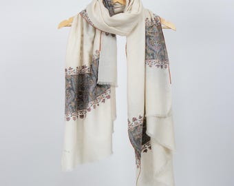 May include: A white pashmina shawl with a paisley pattern border. The shawl is folded and hanging on a wooden hanger.