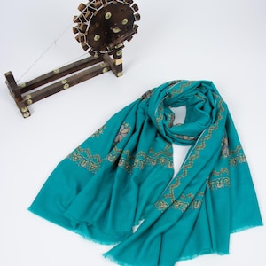 May include: A teal scarf with intricate gold embroidery and a fringed edge is draped on a white surface. A wooden spinning wheel with decorative brass accents sits nearby. The scarf's design features floral and geometric patterns.