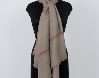 May include: A taupe-colored scarf featuring a delicate, narrow band of intricate embroidery in shades of red, orange, and gold. The scarf has a soft, textured appearance with frayed edges at the bottom, draped elegantly around a mannequin neck.