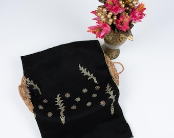 May include: A black scarf with gold, green, and red floral embroidery, displayed on a wicker basket. A vase of pink flowers with gold detailing is also visible.