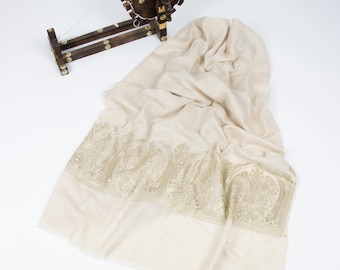 May include: A cream-colored shawl featuring intricate gold-toned embroidery along the border, displayed alongside a small, dark brown wooden spinning wheel with metallic accents and a thin white thread wound around its circular frame.