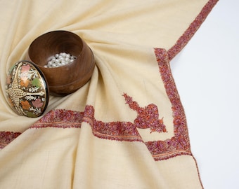 May include: A cream-colored shawl with intricate red embroidery around the edges. A small wooden box with a floral design and white beads inside sits on the shawl.