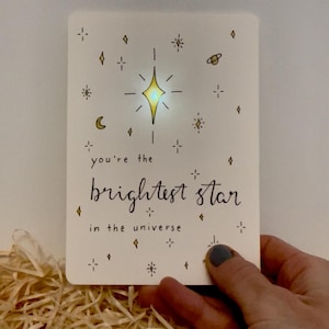 May include: A white card with a glowing yellow star and the handwritten text "you're the brightest star in the universe." The card features small star and diamond illustrations. The card is held over a bed of straw.