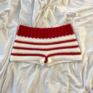 以下が含まれることがあります： 赤と白のストライプ柄のかぎ針編みショートパンツ。ショートパンツは、幅広の赤いウエストバンドと、赤と白の水平ストライプが交互に配置されています。この衣服は手作りで、柔らかい糸で作られているようです。