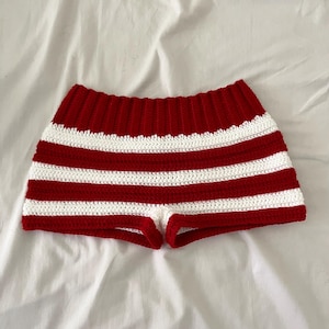 Può includere: Pantaloncini all'uncinetto a righe rosse e bianche. I pantaloncini presentano una serie di bande orizzontali rosse e bianche. La cintura è di colore rosso pieno. Il capo è realizzato con un filato morbido e strutturato.