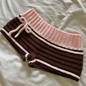 Pode incluir: Calções de crochê marrons e rosa. Os calções apresentam listras horizontais marrons e rosas, com acabamento branco. Uma fita rosa está amarrada na frente. Os calções são feitos à mão e parecem macios e confortáveis.