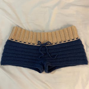 Può includere: Pantaloncini all'uncinetto blu navy con fascia in vita beige. I pantaloncini presentano una chiusura con coulisse e un motivo a punto strutturato. L'indumento è esposto su una superficie bianca, mostrando i dettagli del design fatto a mano.