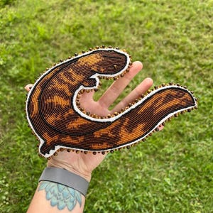 May include: A beaded depiction of a brown and black salamander, curled in a C shape. The salamander is made with brown, black, and white beads.