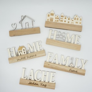 May include: A collection of wooden home decor items. Includes signs with the words "HOME", "FAMILY", and "LACHE", along with house and heart designs. The signs are made of light wood and have a natural finish.