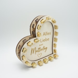 Könnte beinhalten: Eine herzförmige Geschenkbox aus Holz mit dem deutschen Text "Alles Liebe zum Muttertag" und kleinen, goldverpackten Pralinen. Die Box hat ein dekoratives, geschichtetes Design und ist aus hellem Holz gefertigt.