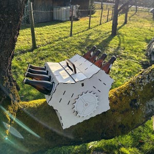 May include: A light-colored wooden beer caddy with a handle, holding six brown beer bottles. The caddy has a gear design on the side and is resting on a tree branch. The background shows a grassy area and trees.