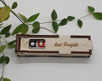Pode incluir: Uma pequena caixa de madeira com uma tampa deslizante. A caixa é gravada com as palavras "du bist perfekt" e dois corações. A caixa é de cor castanho claro.