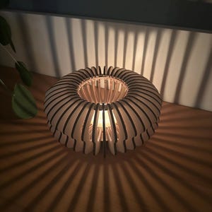 Könnte beinhalten: Eine runde Holzlampe mit einem geschichteten Design. Die Lampe hat eine Lichtquelle im Inneren, die Schatten auf die darunter liegende Oberfläche wirft. Das Design der Lampe weist abwechselnd helle und dunkle Holzstreifen auf.