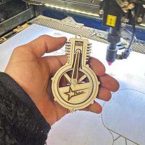 Puede incluir: Un recorte de madera de un motor estilizado con una estrella y la palabra "Peter" inscrita en él. El recorte se está sosteniendo en una mano.