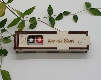 Pode incluir: Uma caixa de madeira com uma barra de chocolate vermelha e branca dentro. A caixa tem as palavras "Du bist die Beste" gravadas, que se traduzem como "Você é a melhor" em português. A caixa é decorada com dois pequenos corações.