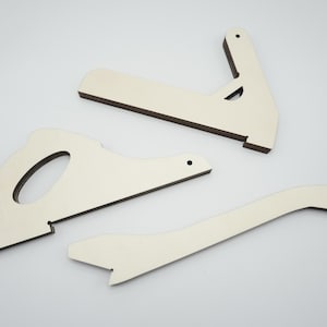 Könnte beinhalten: Ein Satz unfertiger Holzbauteile, darunter ein sägeförmiges Element, ein gebogenes Teil und eine rechtwinklige Stütze. Die Teile haben eine hellbeige Oberfläche mit dunkelbraunen Kanten, ideal für Bastelprojekte.