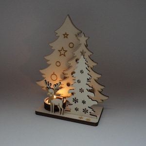 Könnte beinhalten: Weihnachtsdekoration aus Holz mit drei stilisierten Weihnachtsbäumen und einem Rentier. Die Bäume haben Stern- und Kreisausschnitte, und das Rentier steht vor einem Kerzenhalter. Die Basis ist rechteckig, und das Design wird von einem warmen Schein beleuchtet.